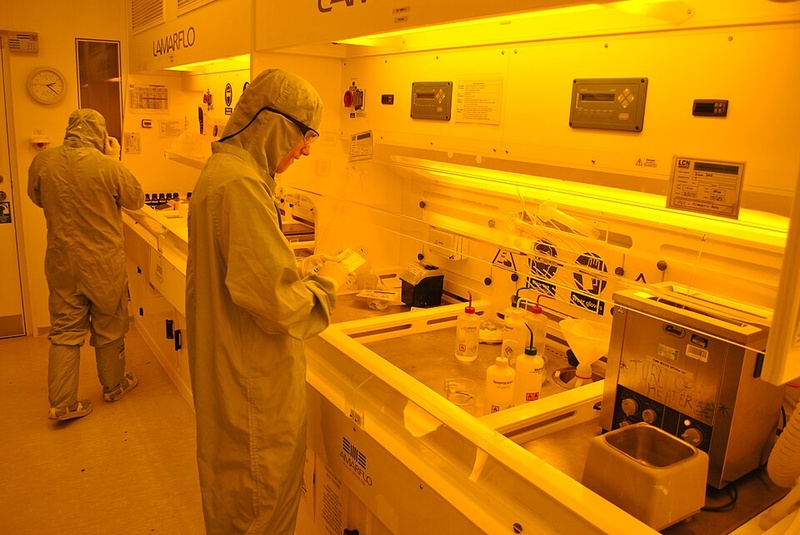 Researchers work in a state-of-the-art photolithography cleanroom at the London Centre for Nanotechnology. The extreme precision required for modern semiconductor manufacturing demands contamination-free environments where even a single dust particle can destroy thousands of transistors. Credit: Wikimedia Commons